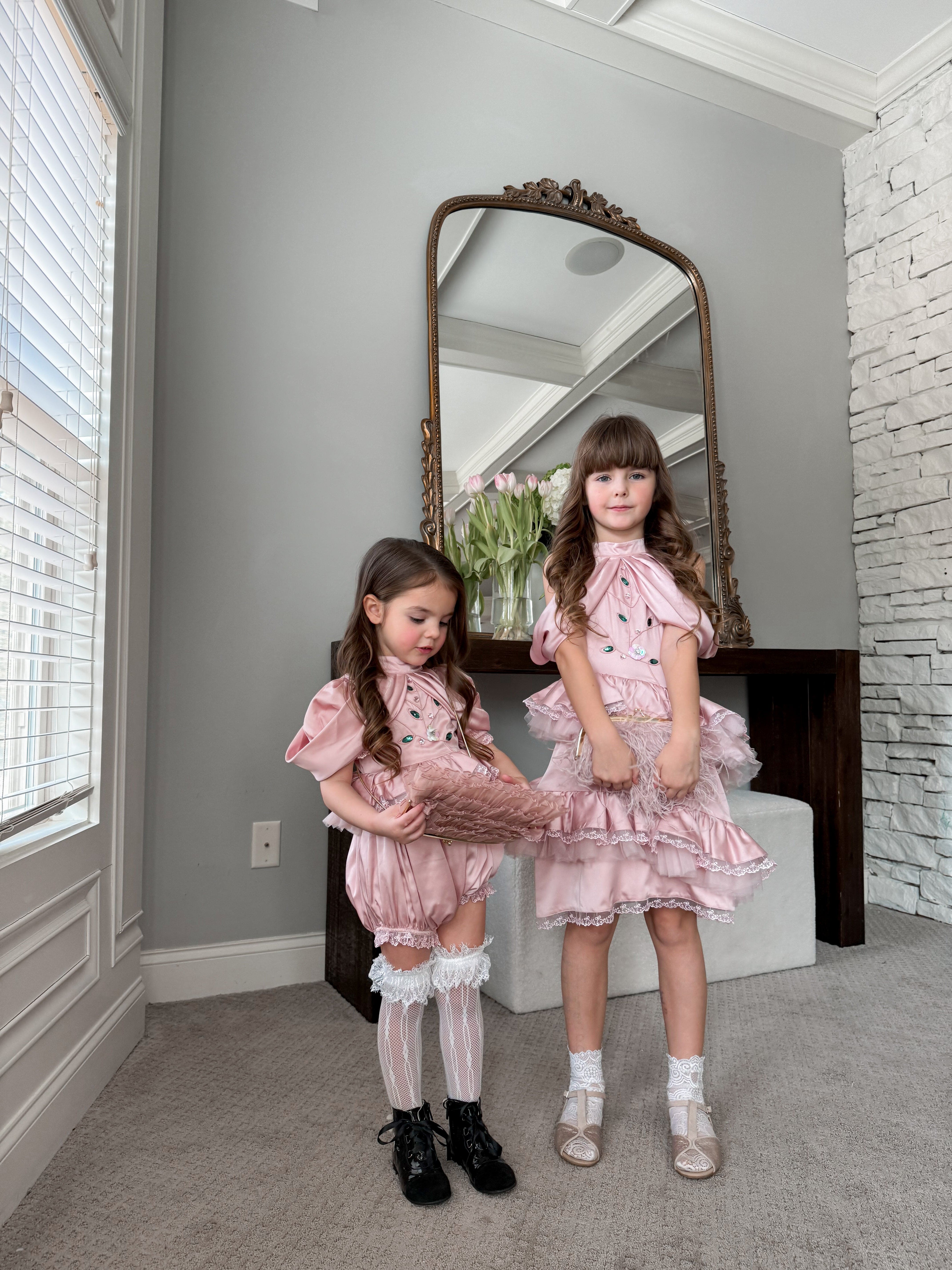 Two girls wearing matching pink satin ballerina dresses with ruffled skirts, photographed in an elegant interior setting