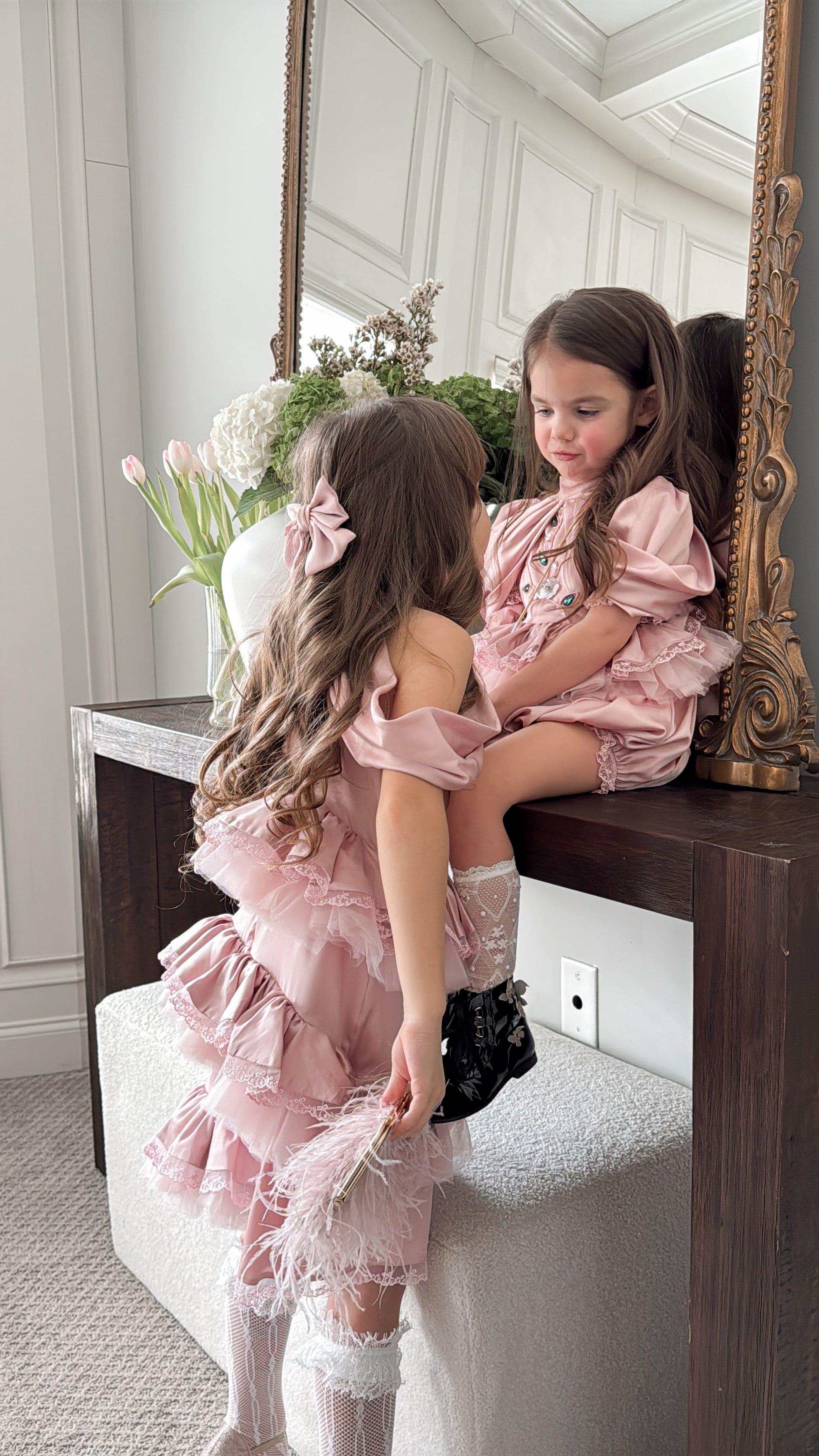 Pink satin ballerina dress styled on a young girl, featuring a ruffled skirt and jewel accented bodice in an elegant mirror setting
