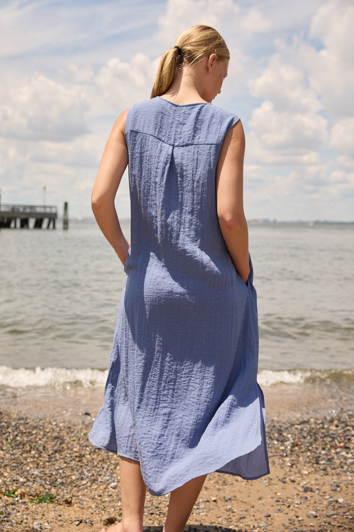 Woman wearing Du & Berry Blue Linen Button Midi Dress standing on a pebbled beach with ocean background