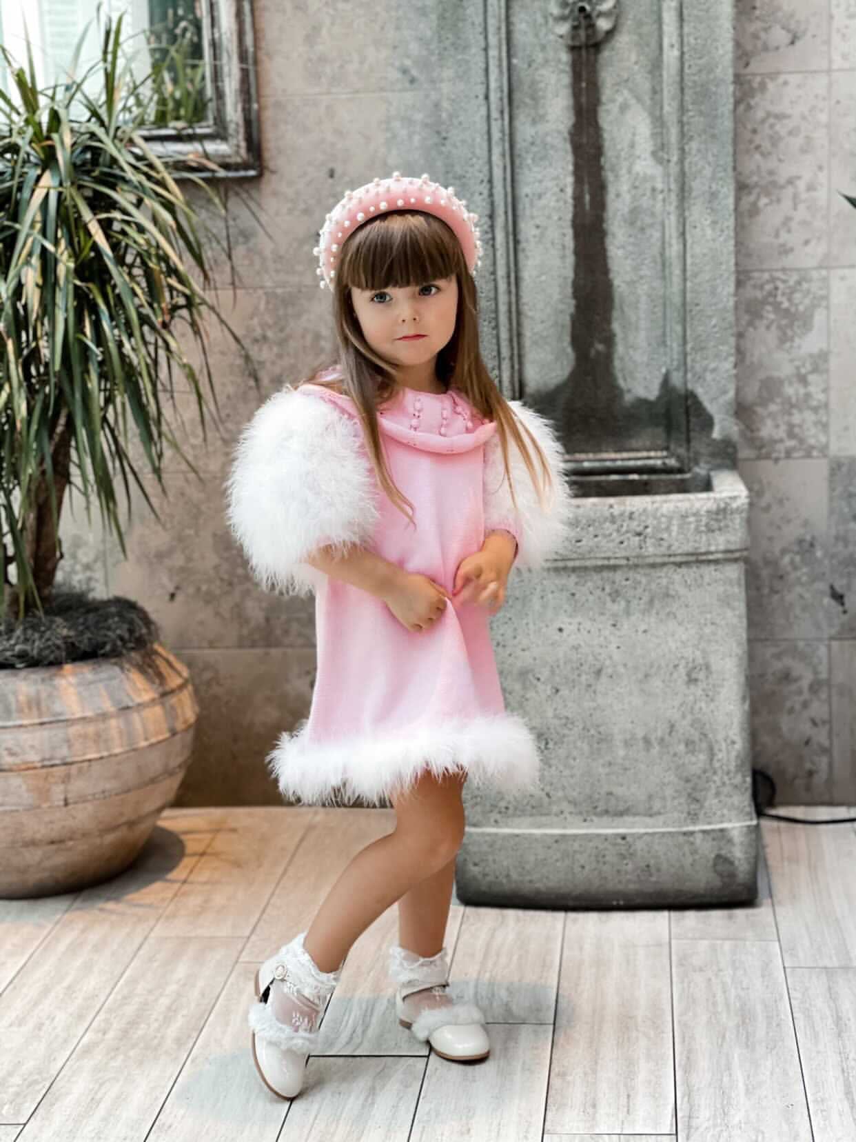 Girl wearing Ella Knit pink feather dress with fluffy sleeves and embellished collar, paired with a pink pearl headband