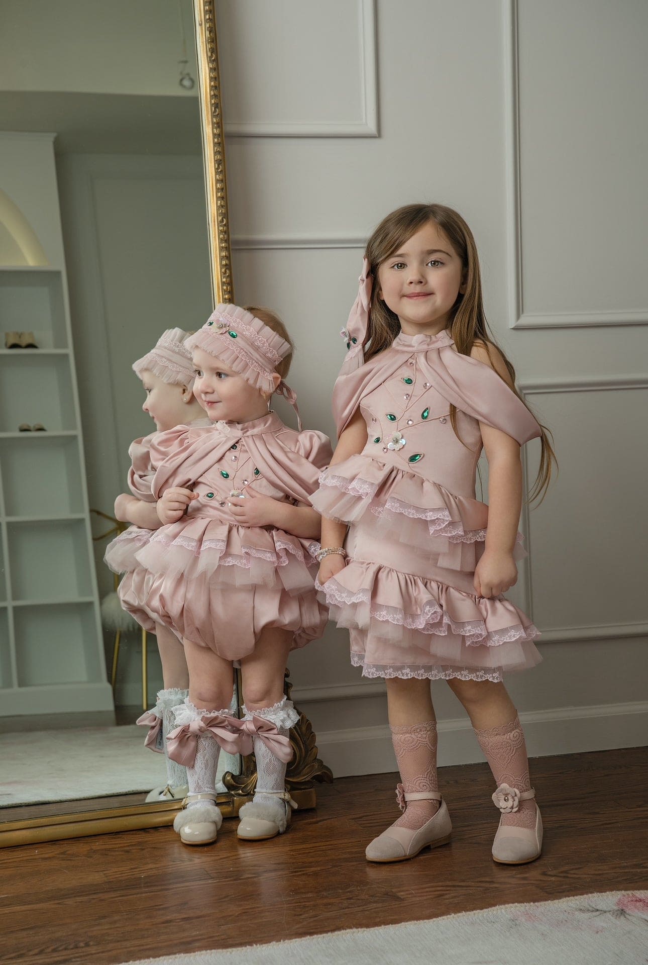 Pink satin ballerina dresses worn by a toddler and young girl, featuring layered ruffles, satin sheen, and jewel accents