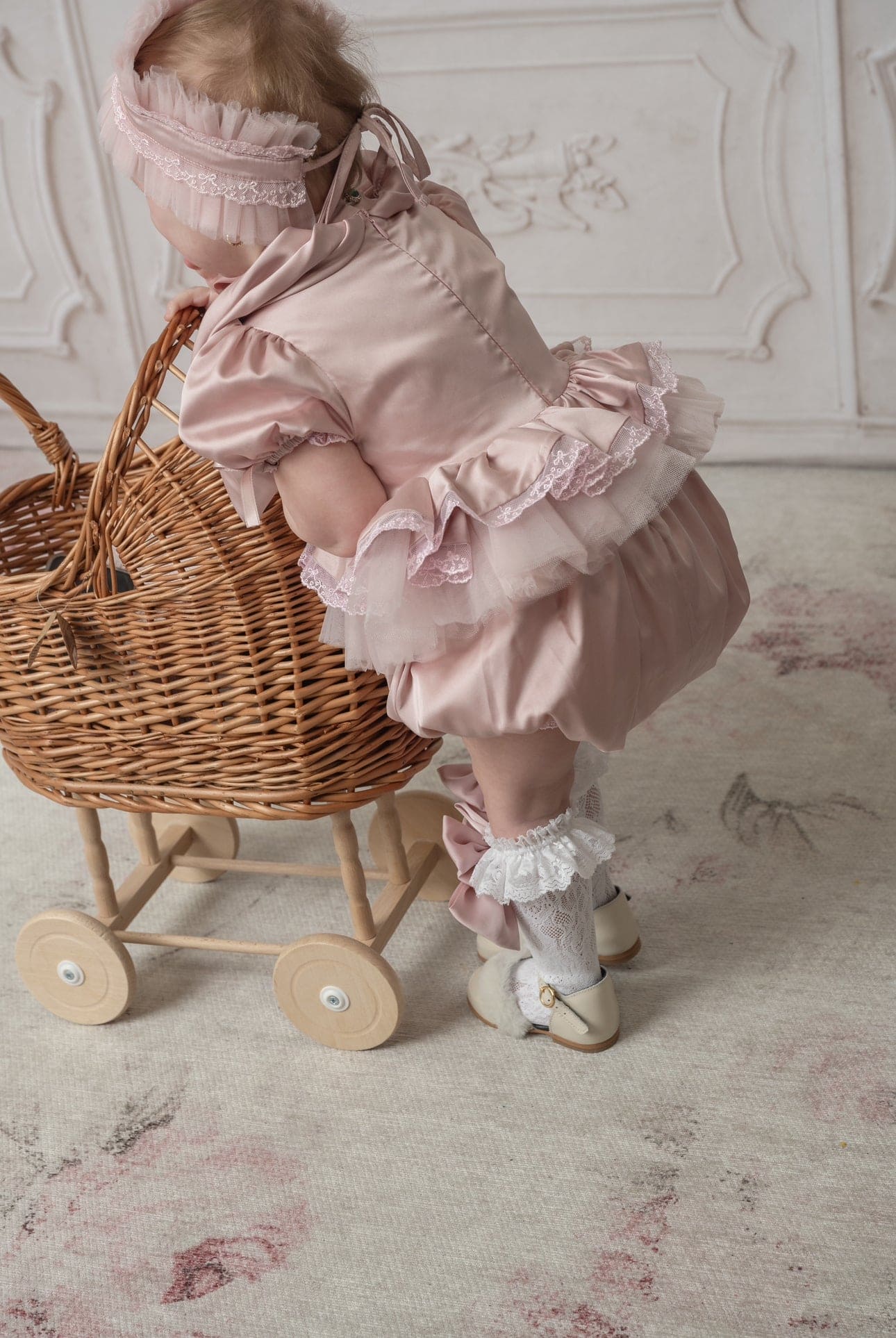 Baby girl in pink satin ruffle romper pushing wicker toy cart